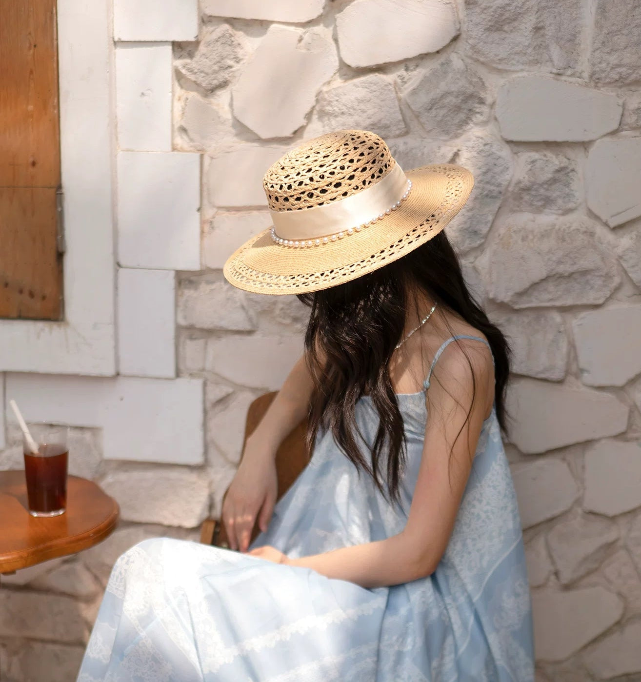 Woman in a light blue dress and straw hat sitting by a stone wall.