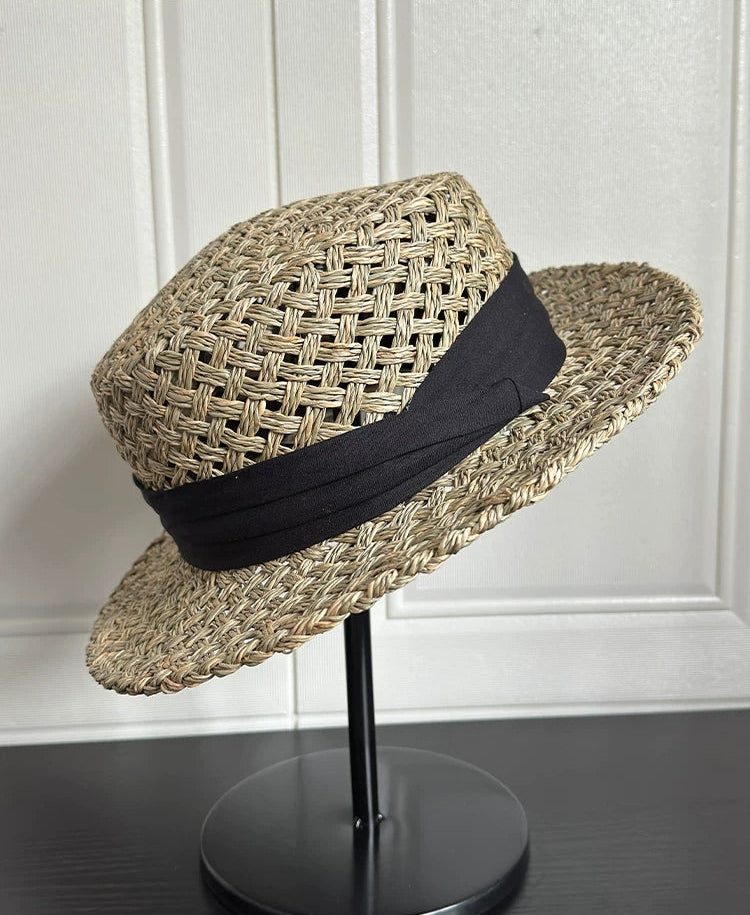 Woven straw hat with a black band on a stand against a white wall.