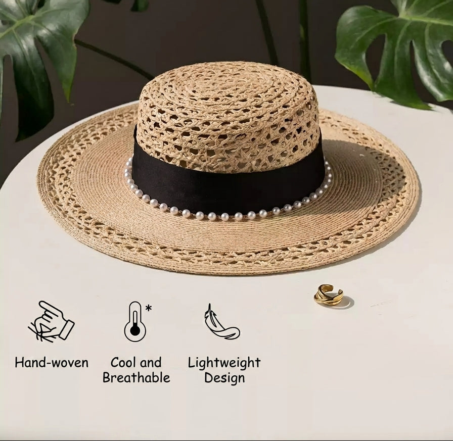 Woven straw hat with black band and pearl details on a white surface with green leaves in the background.